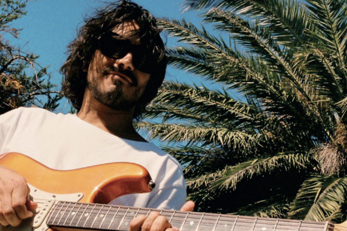Musician Josh Morphett wearing sunglasses plays an electric guitar outdoors under a sunny blue sky with palm trees in the background.